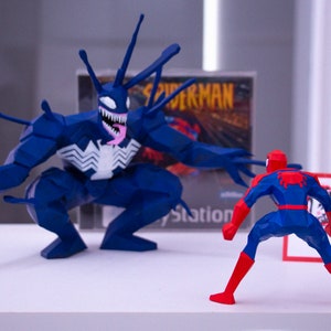 May include: A blue and red Spider-Man figure confronts a large blue Venom figure. The figures are in front of a PlayStation game case. A small red and white sign is to the right of Spider-Man.