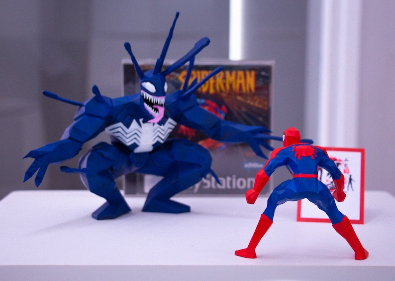 May include: A blue and red Spider-Man figure confronts a large blue Venom figure. The figures are in front of a PlayStation game case. A small red and white sign is to the right of Spider-Man.