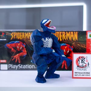 Puede incluir: Figura azul de Venom con detalles blancos, posando frente a una caja de juego de Spider-Man para PlayStation y una caja de Nintendo 64. Tambi&eacute;n hay una peque&ntilde;a tarjeta con texto japon&eacute;s y "MUGICO FIGURES".