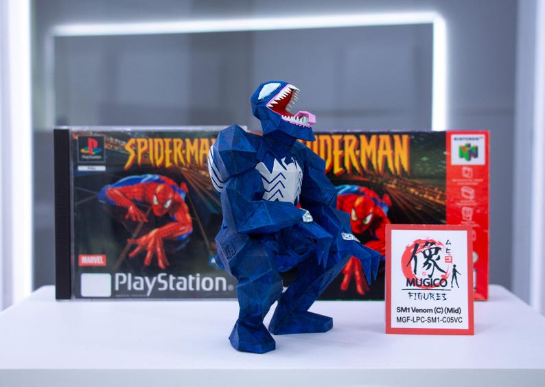 Puede incluir: Figura azul de Venom con detalles blancos, posando frente a una caja de juego de Spider-Man para PlayStation y una caja de Nintendo 64. Tambi&eacute;n hay una peque&ntilde;a tarjeta con texto japon&eacute;s y "MUGICO FIGURES".