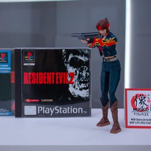 Puede incluir: Una figura de acci&oacute;n roja y negra de una mujer sosteniendo un arma. La figura est&aacute; de pie frente a una caja de videojuego para Resident Evil 2 para Playstation. La caja tiene un zombi en la portada. La figura est&aacute; etiquetada "RE2 Claire (3-4) (Big) MGF-LPC-RE2-C02C3H".
