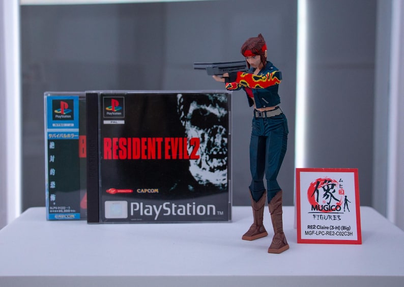 Puede incluir: Una figura de acci&oacute;n roja y negra de una mujer sosteniendo un arma. La figura est&aacute; de pie frente a una caja de videojuego para Resident Evil 2 para Playstation. La caja tiene un zombi en la portada. La figura est&aacute; etiquetada "RE2 Claire (3-4) (Big) MGF-LPC-RE2-C02C3H".