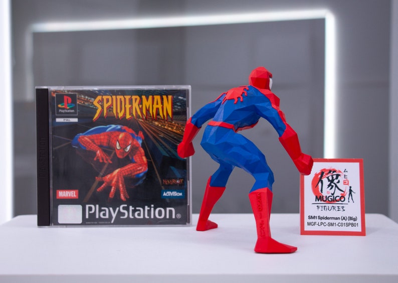 May include: A red and blue 3D printed figurine of Spiderman, standing with one leg forward and holding a red and white card with Japanese text and the words "MUGICO FIGURES" and "SM1 Spiderman (A) (Big) MGF-LPC-SM1-C015PB01". The figurine is next to a Playstation game case for Spiderman.
