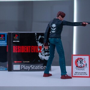 May include: A video game case for Resident Evil 2 for the Playstation console. The case is blue and white with the game title in red. A figure of a character from the game is standing in front of the case. The figure is wearing a black jacket with a skull on the back, blue jeans, and red shoes. The figure is holding a gun in their right hand. A red and white card with Japanese text is in the foreground.