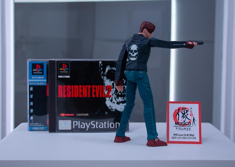 May include: A video game case for Resident Evil 2 for the Playstation console. The case is blue and white with the game title in red. A figure of a character from the game is standing in front of the case. The figure is wearing a black jacket with a skull on the back, blue jeans, and red shoes. The figure is holding a gun in their right hand. A red and white card with Japanese text is in the foreground.