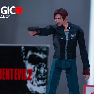 May include: A black and blue action figure of a man holding a gun. The figure is wearing a black jacket, blue jeans, and a black shirt. The figure is standing in front of a video game case with the text "RESIDENT EVIL 2" on it.