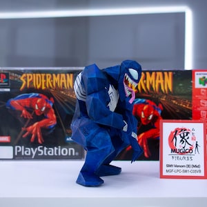 May include: A blue and white Venom figure is positioned in front of a PlayStation Spider-Man game box and a Nintendo 64 game box. A small red and white sign reads "MUGICO FIGURES" and "SM1 Venom (B) (Mid)".