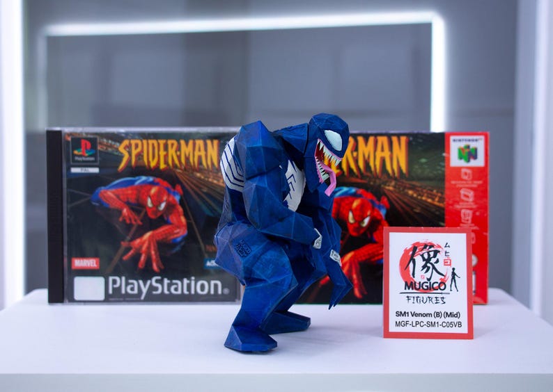 May include: A blue and white Venom figure is positioned in front of a PlayStation Spider-Man game box and a Nintendo 64 game box. A small red and white sign reads "MUGICO FIGURES" and "SM1 Venom (B) (Mid)".