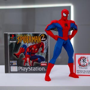 May include: A red and blue Spider-Man figure stands next to a PlayStation game case for Spider-Man 2. The figure has a geometric design. A small sign with Japanese text is also present.