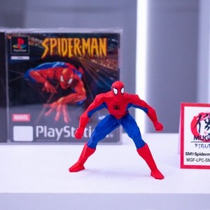 May include: A red, blue, and black Spider-Man action figure stands before a PlayStation game case. The game case features the title "SPIDERMAN" and the Marvel logo. A small card with Japanese text is also present.
