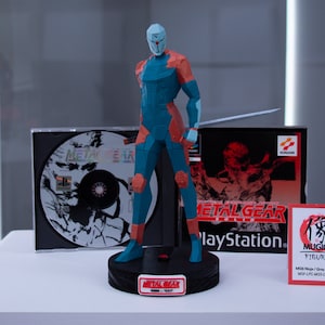 Puede incluir: Una figura poligonal azul y naranja de un ninja con una espada, basada en la serie de videojuegos Metal Gear. La figura está sobre una base negra, con un disco de juego y una caja en el fondo. La caja dice "Metal Gear" y "PlayStation".