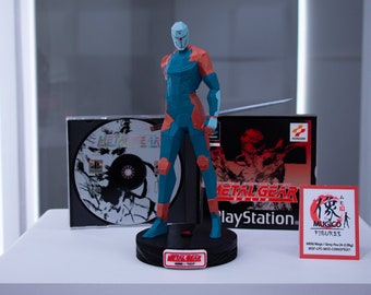 May include: A blue and orange polygonal figure of a ninja with a sword, based on the Metal Gear video game series. The figure stands on a black base, with a game disc and box in the background. The box reads "Metal Gear" and "PlayStation".