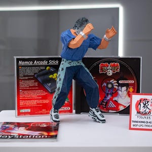 Può includere: Una action figure di Tekken 3 in posa da combattimento, con un completo blu e verde acqua, si trova di fronte a una scatola Namco Arcade Stick, un disco di gioco di Tekken 3 e una custodia di gioco per PlayStation. È presente anche una piccola carta rossa con testo giapponese.