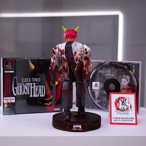 Peut inclure: Une figurine peinte en rouge et blanc représentant un homme en costume portant un masque de démon rouge. La figurine est posée sur un socle en bois avec une étiquette noire indiquant "Clock Tower Ghost Head". La figurine est exposée à côté d'un boîtier de jeu vidéo pour "Clock Tower: Ghost Head" et d'un disque de jeu vidéo. Une pancarte rouge et blanche indique "MUGICO FIGURES CT GHOST HEAD (JP)  MUGI-JP-CTGHOS-011MA"