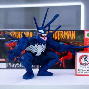May include: A blue Venom figure with a white spider emblem on its chest, posed in front of a Spider-Man PlayStation game box. The figure has a white mouth with sharp teeth and a pink tongue. A red sign with Japanese text is also present.