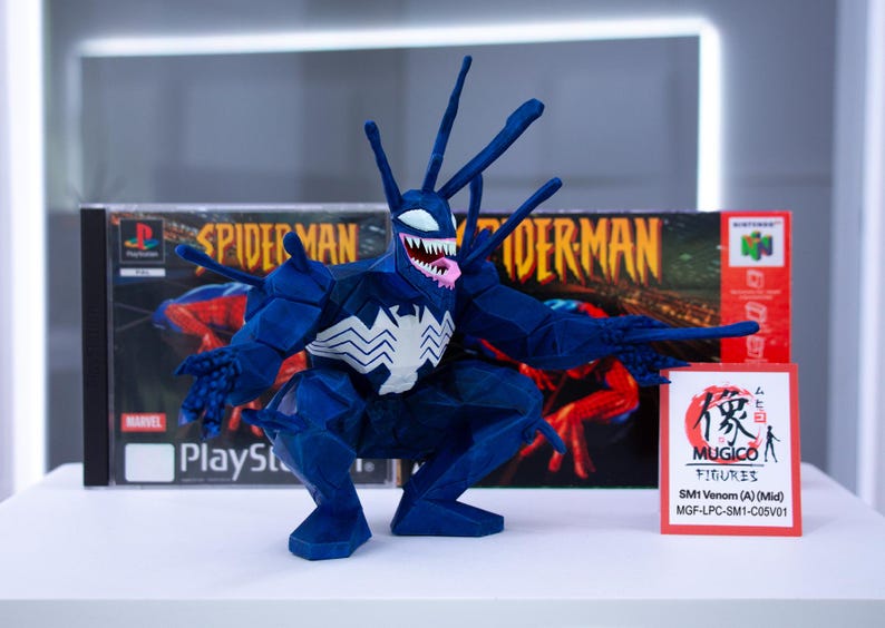 May include: A blue Venom figure with a white spider emblem on its chest, posed in front of a Spider-Man PlayStation game box. The figure has a white mouth with sharp teeth and a pink tongue. A red sign with Japanese text is also present.