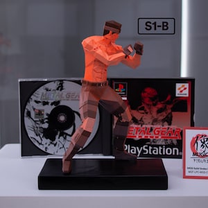 Könnte beinhalten: Eine rote und braune Polygon-Figur von Solid Snake aus dem Videospiel Metal Gear Solid. Die Figur ist in Kampfstellung und steht auf einem schwarzen Sockel. Die Figur steht neben einem Metal Gear Solid PlayStation-Spielecase und einer kleinen roten Karte mit dem Text "MUGICO FIGURES" und "MOS Solid Snake 2014-01 (Big) MOF-LPC-MOS-C3185501".