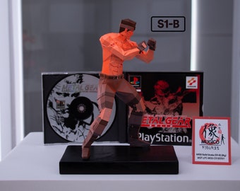 May include: A red and brown polygon-style figurine of Solid Snake from the Metal Gear Solid video game. The figurine is posed in a fighting stance and is standing on a black base. The figurine is next to a Metal Gear Solid PlayStation game case and a small red card with the text "MUGICO FIGURES" and "MOS Solid Snake 2014-01 (Big) MOF-LPC-MOS-C3185501".