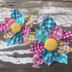 May include: Two colourful gingham fabric flower hair clips. Each flower is made of five petals in different colours, including pink, blue, yellow, and green. The centre of each flower has a yellow button.