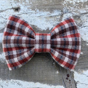May include: A brown, white, and red plaid bow tie. The bow tie is made of a soft, textured fabric.