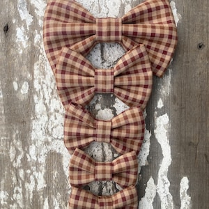 May include: A collection of five brown and burgundy plaid bow ties of varying sizes, arranged vertically. The bow ties are made of fabric with a classic gingham pattern. The background is a weathered, light-colored wooden surface.
