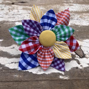 May include: A colourful gingham flower pin with a yellow centre. The flower is made of fabric and has a variety of colours, including red, blue, green, yellow, and purple.