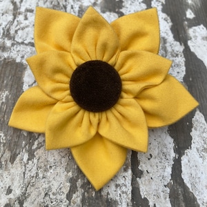May include: A yellow fabric sunflower with a brown felt center. The sunflower is on a rustic wooden background.