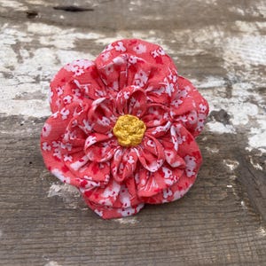 May include: A fabric flower in coral with a white floral print, featuring a yellow centre. The flower is made of layered petals and rests on a weathered wooden surface.