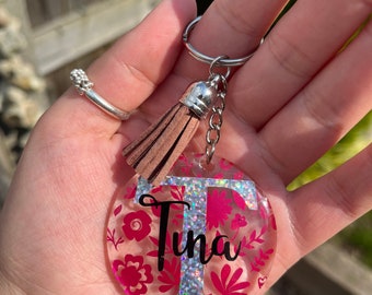 May include: A clear round keyring with a pink floral design and the name "Tina" in black lettering. The keyring has a silver tassel.