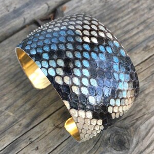 Könnte beinhalten: Ein breites Armband aus blauem und schwarzem Schlangenleder mit einem goldenen Metallband.