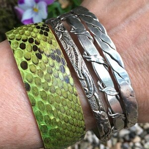 Pu&ograve; includere: Un set di tre braccialetti d'argento, uno con un designo intrecciato, uno con un designo floreale e uno con una superficie liscia. I braccialetti sono indossati al polso di una persona, insieme a un braccialetto di pelle di serpente verde.