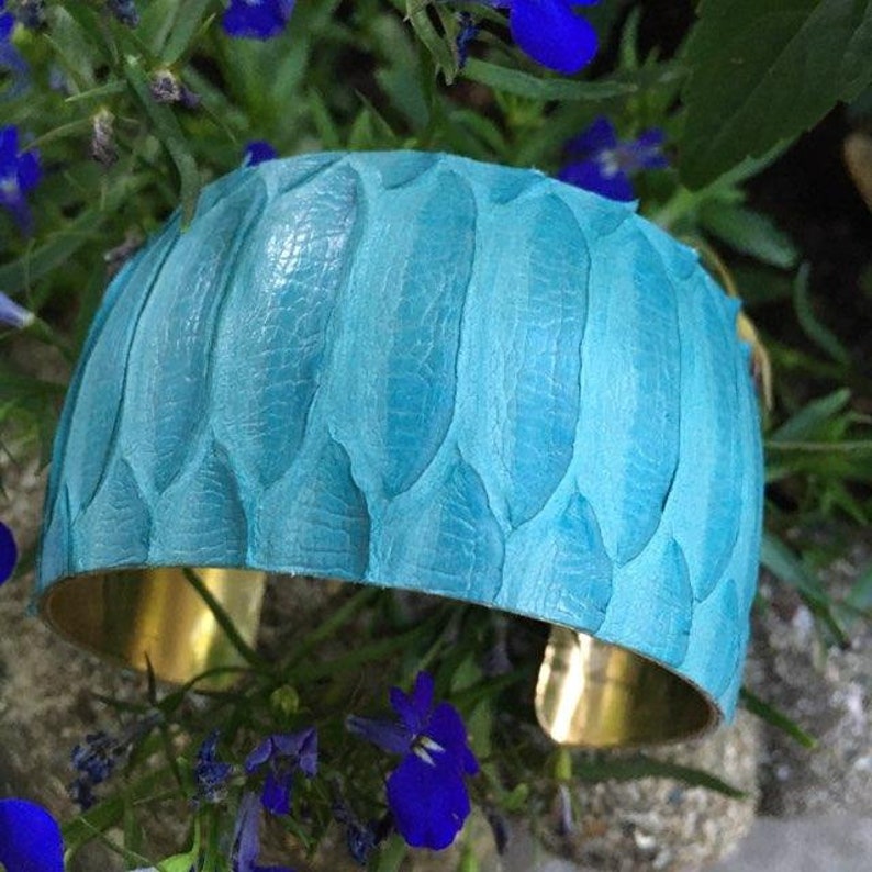 Pode incluir: Um bracelete de couro azul turquesa com um padr&atilde;o texturizado de escamas. O bracelete &eacute; feito de lat&atilde;o e tem um design largo e plano.