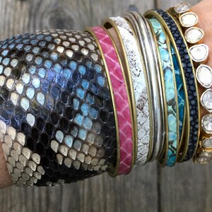 May include: A stack of colourful bracelets, including a snakeskin bracelet, a pink leather bracelet, a white leather bracelet, a silver bracelet, a turquoise leather bracelet, and a black leather bracelet with small white stones.