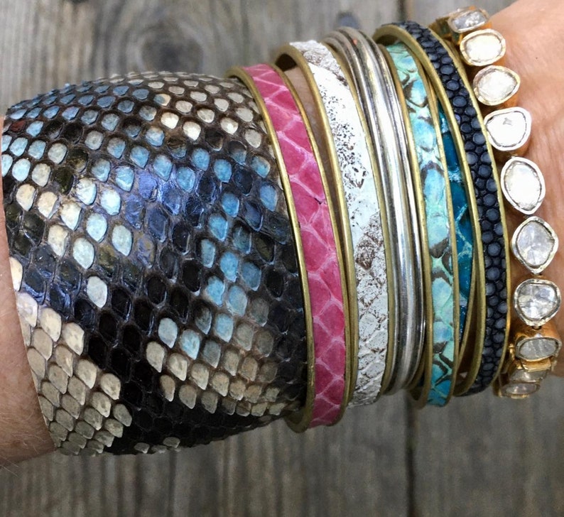 May include: A stack of colourful bracelets, including a snakeskin bracelet, a pink leather bracelet, a white leather bracelet, a silver bracelet, a turquoise leather bracelet, and a black leather bracelet with small white stones.