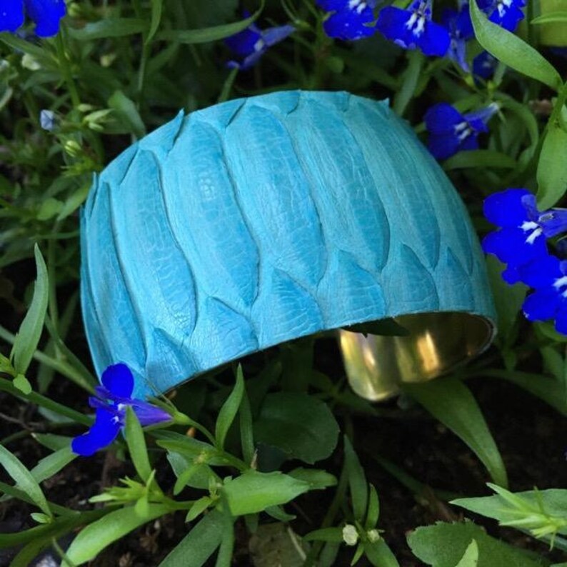 Pode incluir: Um bracelete de couro azul turquesa com um padr&atilde;o texturizado em forma de folha. O bracelete tem uma base de metal dourado.