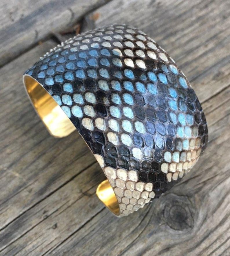 May include: A wide cuff bracelet made of blue and black snakeskin with a gold metal band.