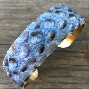 K&ouml;nnte beinhalten: Ein blaues und wei&szlig;es Schlangenleder-Armband mit einem Messingband. Das Armband ist aus echtem Schlangenleder gefertigt und hat ein einzigartiges, strukturiertes Muster.