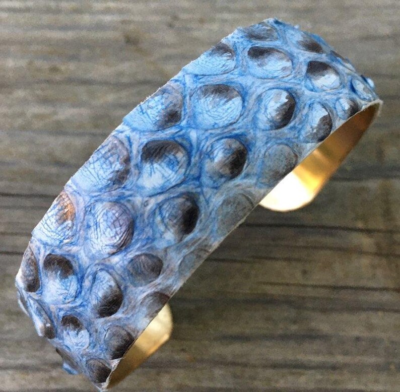 K&ouml;nnte beinhalten: Ein blaues und wei&szlig;es Schlangenleder-Armband mit einem Messingband. Das Armband ist aus echtem Schlangenleder gefertigt und hat ein einzigartiges, strukturiertes Muster.