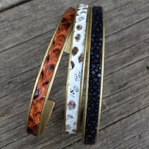 Könnte beinhalten: Drei Lederarmbänder mit goldenen Metallakzenten. Die Armbänder sind übereinander gestapelt. Das Leder ist in drei verschiedenen Mustern: orange, weiß mit braunen Flecken und schwarz.