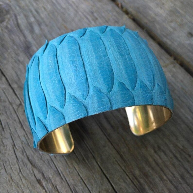 Pode incluir: Um bracelete de couro azul turquesa com base de lat&atilde;o. O couro tem um padr&atilde;o texturizado que se assemelha a escamas.