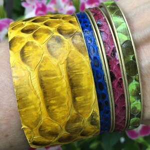May include: A set of four colorful snake skin bracelets. The bracelets are stacked on top of each other, with the yellow bracelet on the bottom, followed by a blue bracelet, a burgundy bracelet, and a green bracelet.