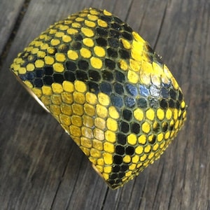 Könnte beinhalten: Ein gelbes und schwarzes Schlangenlederarmband. Das Armband ist aus echtem Schlangenleder gefertigt und hat ein einzigartiges Muster.