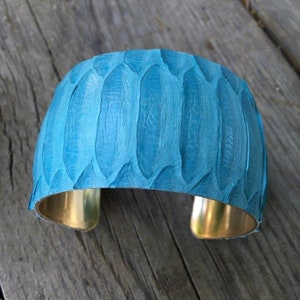 Pode incluir: Um bracelete largo de couro turquesa com uma banda de metal dourado. O couro tem um padr&atilde;o texturizado.