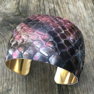 Könnte beinhalten: Ein breites goldenes Armreif mit einem Schlangenhautmuster in den Farben Rosa, Schwarz und Grau.