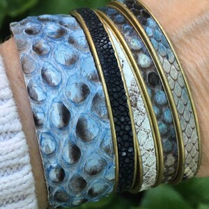 K&ouml;nnte beinhalten: Stapel von vier Lederarmb&auml;ndern mit einem blauen, wei&szlig;en und schwarzen Muster. Die Armb&auml;nder sind aus Schlangenleder und haben einen Messingbesatz.
