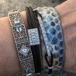 Pu&ograve; includere: Un set di quattro braccialetti, tra cui un delicato braccialetto d'argento con un design geometrico e pietre preziose trasparenti, un braccialetto in cavo intrecciato nero, un braccialetto d'argento con una fila di pietre preziose trasparenti e un braccialetto in pelle con motivo blu e bianco.