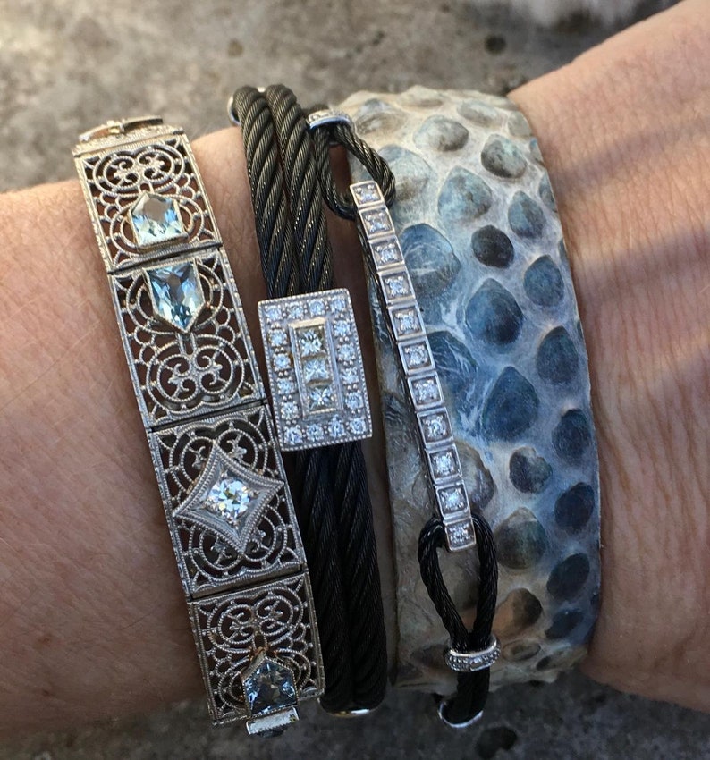 Pu&ograve; includere: Un set di quattro braccialetti, tra cui un delicato braccialetto d'argento con un design geometrico e pietre preziose trasparenti, un braccialetto in cavo intrecciato nero, un braccialetto d'argento con una fila di pietre preziose trasparenti e un braccialetto in pelle con motivo blu e bianco.