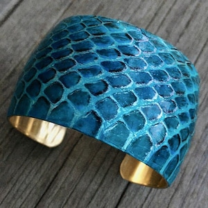 Könnte beinhalten: Ein breites, türkisblaues Armreif mit Schlangenhautmuster. Das Armband ist aus Metall und hat eine goldene Oberfläche.