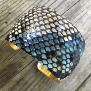 May include: A wide, blue and black snakeskin cuff bracelet with a gold metal band. The bracelet is made of real snakeskin and has a unique, textured design.