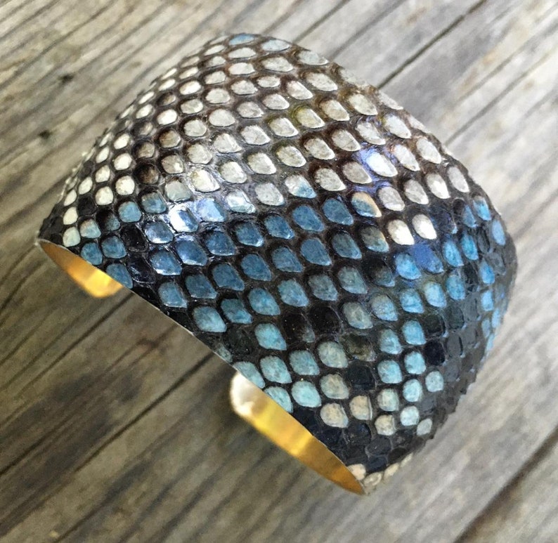 May include: A wide, blue and black snakeskin cuff bracelet with a gold metal band. The bracelet is made of real snakeskin and has a unique, textured design.